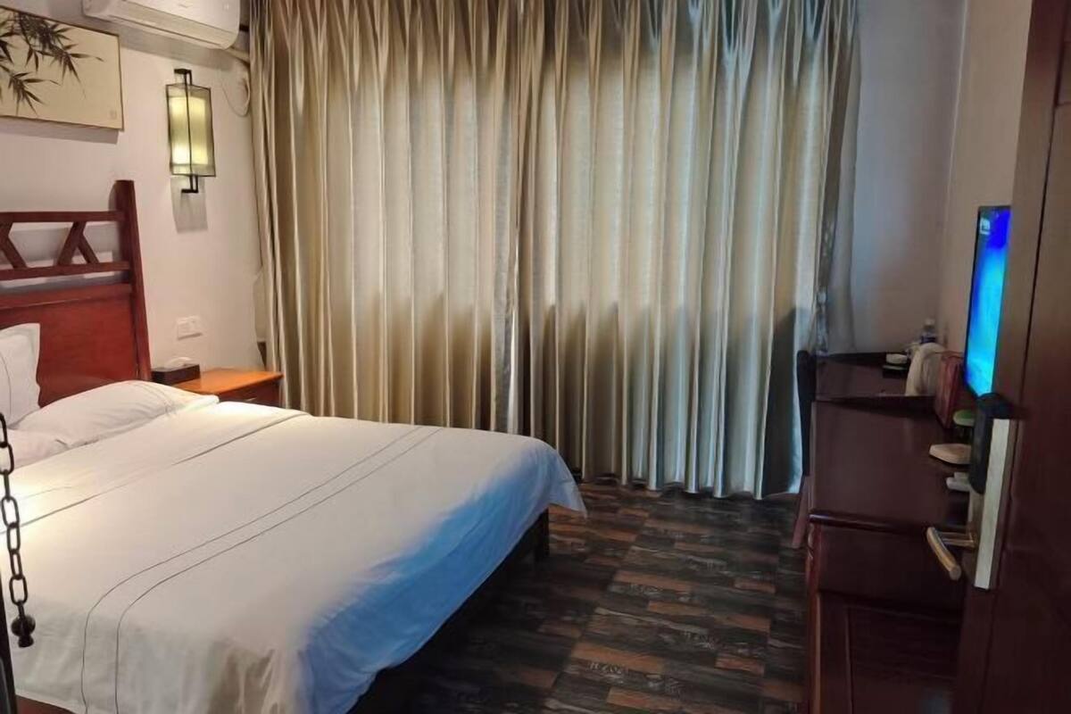 A hotel room with a bed, curtains, a desk with a computer, and a wall-mounted lamp.