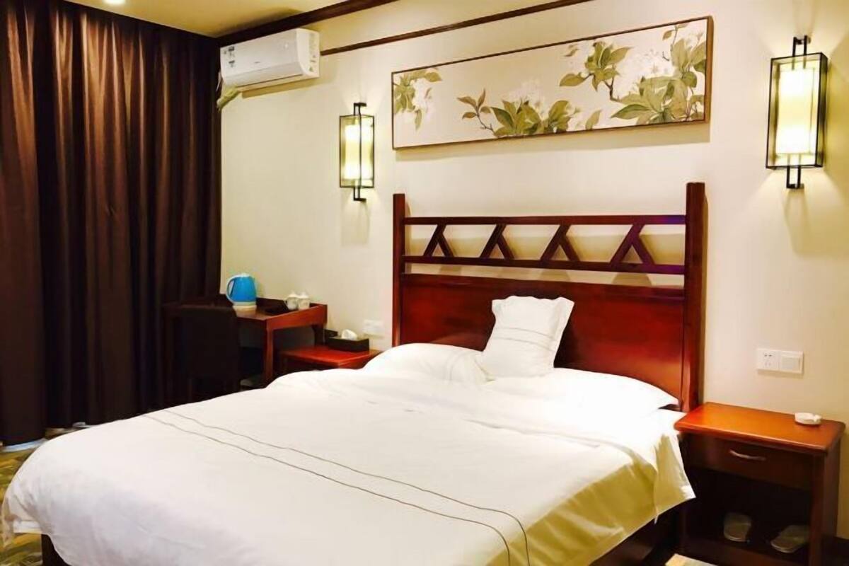 A hotel room with a bed, bedside tables, a desk, a lamp, and a wall-mounted air conditioner.