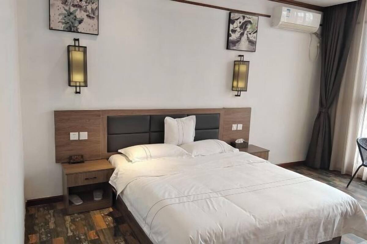 A hotel room with a bed, bedside tables, wall-mounted lights, artwork, and an air conditioning unit.