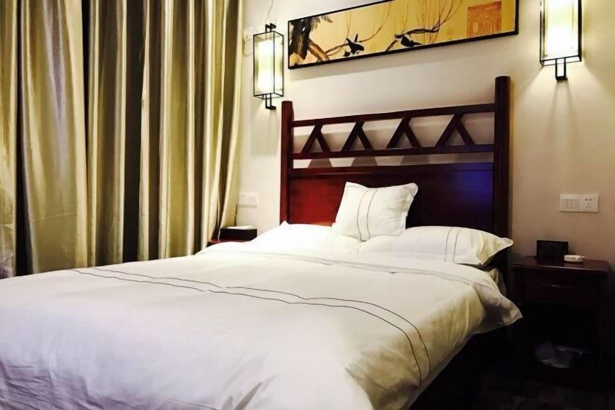 A hotel room with a large bed, a wooden headboard, two bedside tables, a wall-mounted lamp, and a painting above the bed.