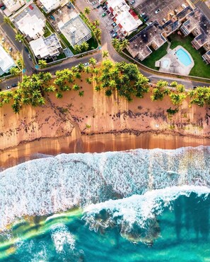 An aerial view of a coastal area with a beach, ocean waves, and a residential area with buildings and parked cars.