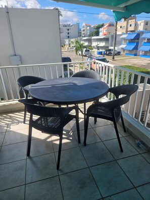 A balcony with a round table and four chairs, a tiled floor, and a view of buildings and parked cars in the background.