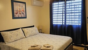 Un llit ben fet amb un edredó estampat i dues tovelloles doblegades, un ventilador de sostre, una finestra amb cortines i un aire condicionat fixat a la paret.