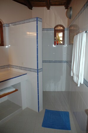 A bathroom with white tiles, a blue-tiled shower area, a wooden shelf, and a blue bath mat.