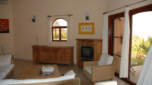 A spacious living room with a wooden cabinet, a television, wicker furniture, and a large window with curtains. - Les Alizés Cap Skirring (Cap Skirring)