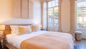 A hotel room with a large bed, two bedside tables, a window with a view of buildings, and a door with a balcony. - Casa Azul - Santa Marinha (Vila Nova de Gaia)