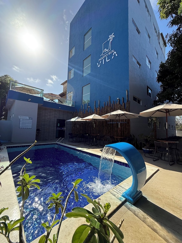 Una piscina con fuente acuática, rodeada de mobiliario exterior y un edificio de fachada azul.