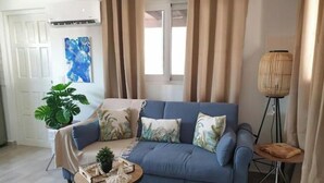 Een woonkamer met een blauwe bank, een klein rond tafeltje met een plant, een grote potplant en een vloerlamp.