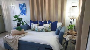 Een moderne slaapkamer met een blauwe slaapbank, een witte dekbedovertrek en meerdere kussens. Op de vloer ligt een klein rond vloerkleed, op de bank staat een pot met een plant en een vloerlamp staat naast het bed.