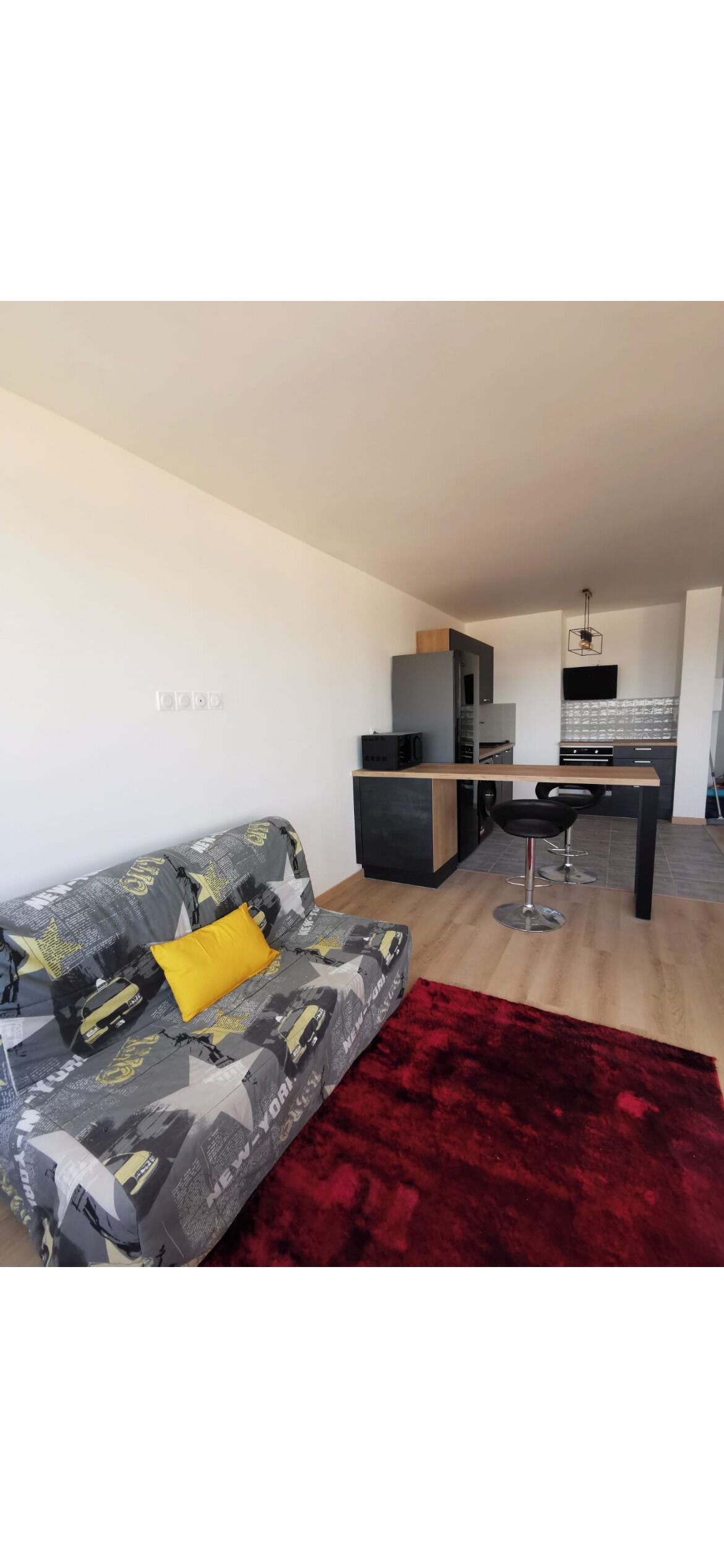 Una sala de estar moderna con un sofá gris, un tapete rojo y una zona de cocina al fondo.