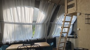 A cozy dining area with a wooden table and chairs, a ladder, and a view of greenery through sheer curtains.