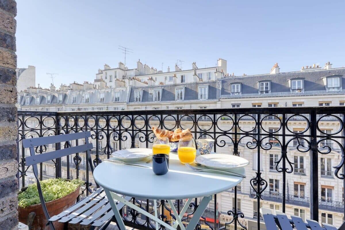 A balcony with a table set for breakfast, including croissants and orange juice, overlooking a row of terraced houses.