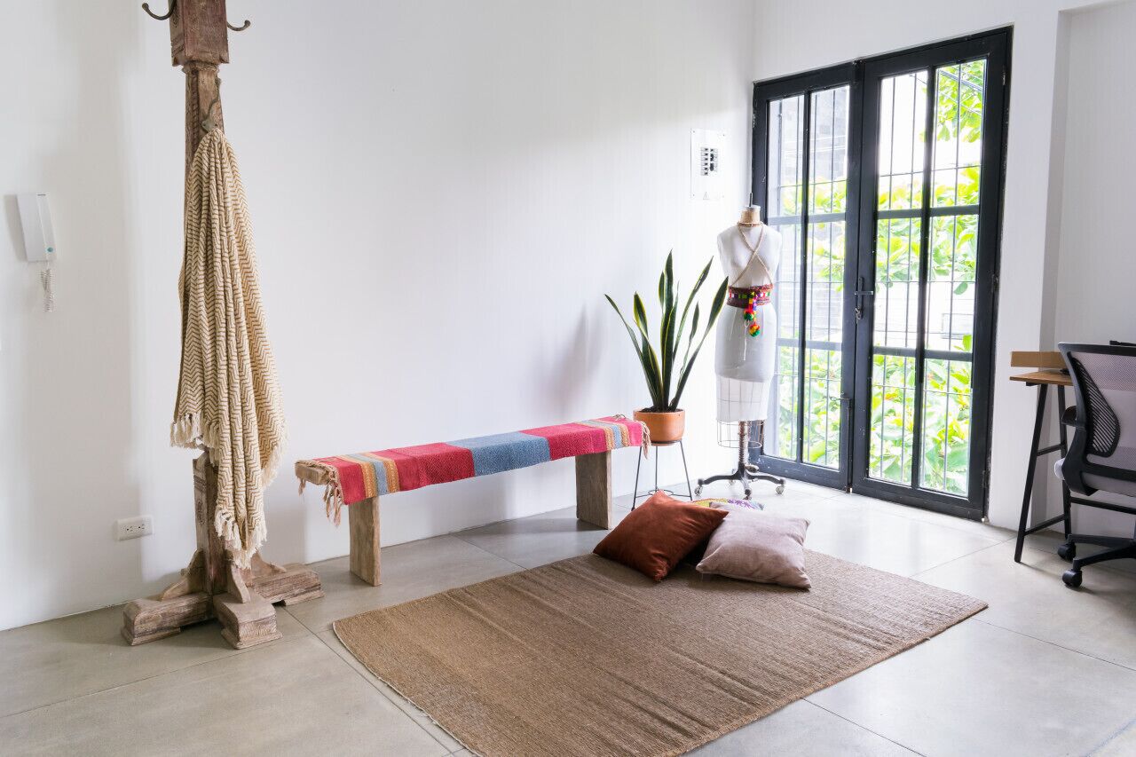 Una habitación con un banco, una cuerda colgante, una planta en maceta, un maniquí y un ventanal grande.