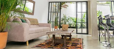 A living room with a sofa, coffee table, and chairs, surrounded by large windows and potted plants.