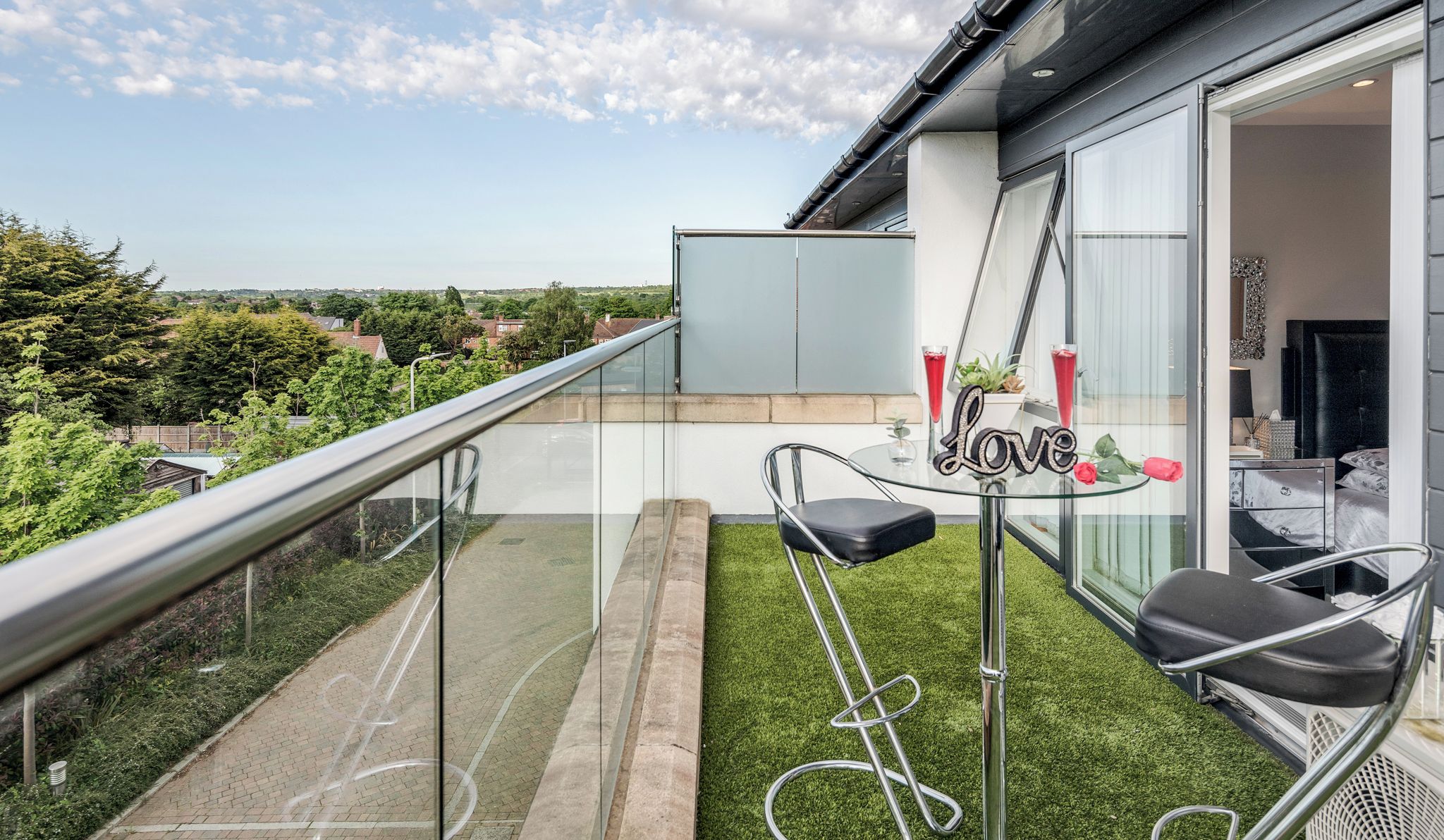 A balcony with a glass railing, a small table with two chairs, and a view of the surrounding area.