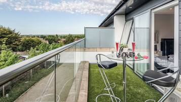 A balcony with a glass railing, a small table with two chairs, and a view of the surrounding area.