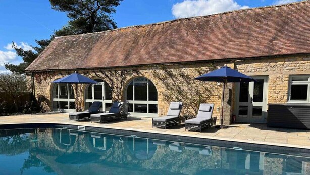 A swimming pool with lounge chairs and umbrellas, a stone building with arched windows, and a clear blue sky. - Luxury Cottage with Pool (Bredon)