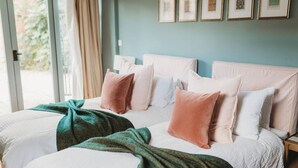 A double bed with multiple pillows and a green blanket, a wooden nightstand, and a window with curtains. - Luxury Cottage with Pool (Bredon)