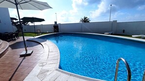 Una piscina con muelle de baldosas, escalera metálica y sombrilla blanca.