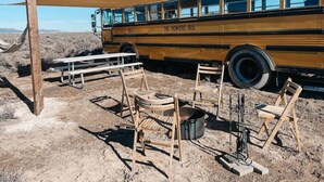 Un ancien autobus scolaire est garé dans un paysage désertique, avec des tables de pique-nique et des chaises en bois à proximité.