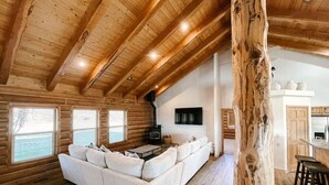 Interior de una cabaña de troncos con un sofá blanco grande, piso de madera, chimenea y una zona de cocina.