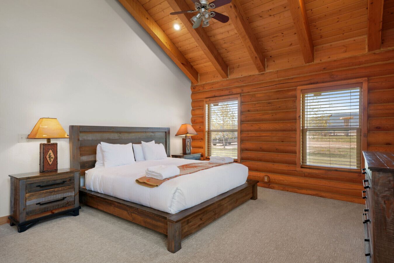 Een slaapkamer in een blokhut met een houten bed, twee nachtkastjes met lampen, een plafondventilator en grote ramen.