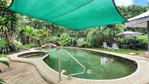 A swimming pool with a green shade sail, surrounded by lush greenery and a patio area with outdoor furniture.