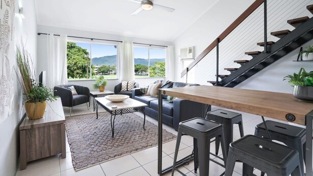 A modern living room with a sofa, coffee table, and bar stools. There is a staircase leading to an upper level.