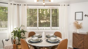 Un comedor con una mesa redonda, sillas, una araña y vistas a árboles a través de grandes ventanales.