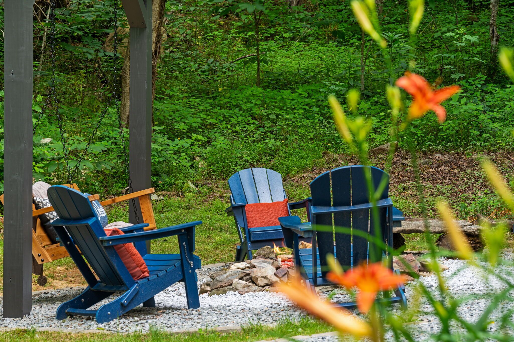 Dos sillas Adirondack con cojines, un brasero y un bosque natural de fondo.