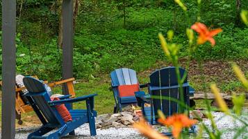 Dos sillas Adirondack con cojines, un brasero y un bosque natural de fondo.