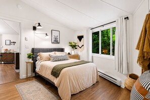 A bedroom with a bed, bedside tables, a nightstand, a window with curtains, a plant, and a hanging light fixture.