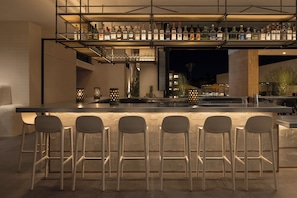 A bar area with a long counter, multiple stools, and a variety of bottles on shelves. - Drift Palm Springs, a Member of Design Hotels (Palm Springs)