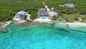 An aerial view of a coastal property with a large house, a swimming pool, and a dock extending into turquoise waters. - Newly Built Luxury 5- bedroom Beachfront Villa with Infinity Heated Pool (Exuma)