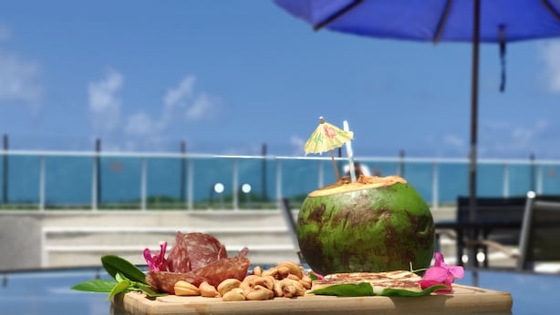 A tropical drink setup with a coconut and a straw, surrounded by snacks and a flower on a wooden board, with a blue umbrella and a view of the sea in the background. - Lindo Apartamento com Varanda e Vista Mar (Barra de São Miguel)