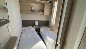 A compact RV bedroom with two beds, a small table, and a shelf with decorative items. - M697 - 8 berth with veranda - Pet Friendly (Camber)