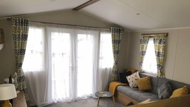 A living room with a sofa, a small table, and a lamp. There are curtains on the windows. - Beechwood 18 -With Veranda -  No pets Allowed (Camber)