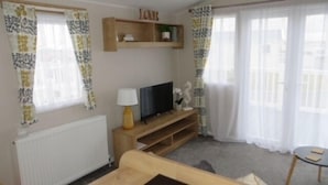 A living room with a TV on a wooden stand, a lamp, and a shelf with decorative items. - Beechwood 18 -With Veranda -  No pets Allowed (Camber)