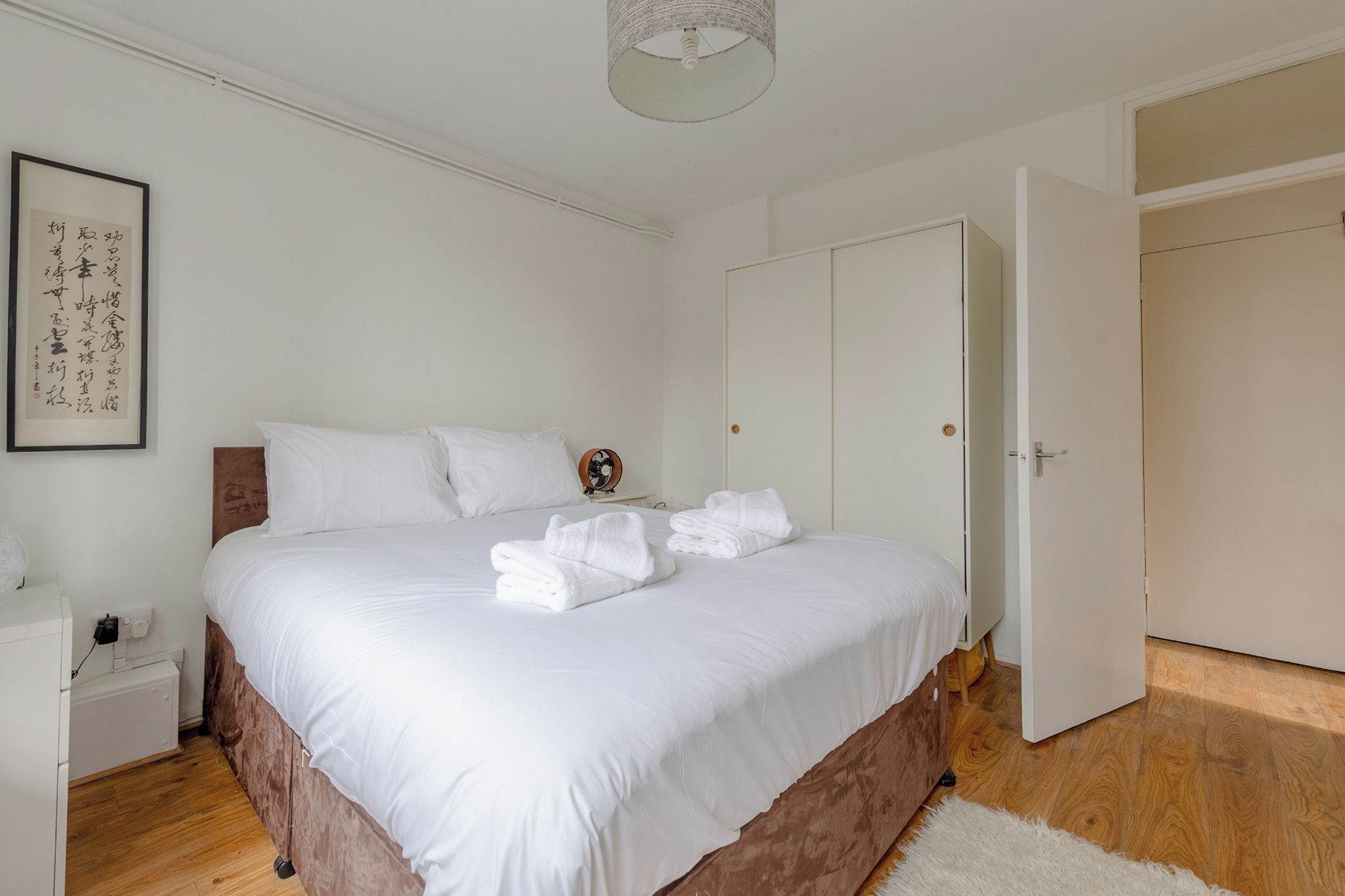 A neatly made bed with white linens and towels, a bedside table with a lamp, and a framed calligraphy piece on the wall.