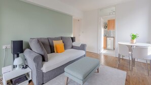 Un salon moderne avec un canapé gris, une petite table, une chaise blanche et un coin cuisine visible à travers une porte ouverte.