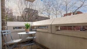 Un petit balcon extérieur avec une table et deux chaises, un pot de fleurs et une vue sur des immeubles et des arbres.