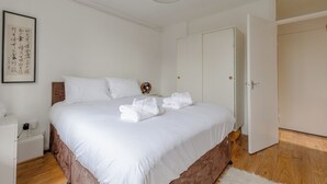 Un lit bien fait, avec du linge de lit et des serviettes blanches, une table de chevet avec une lampe, une œuvre d’art encadrée au mur et un plancher en bois.