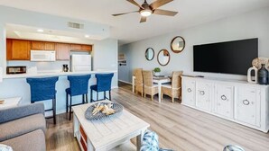 A modern living room with a dining area, kitchen, and a TV mounted on the wall. - Destin West - Sandpiper 407 (Okaloosa Island)