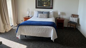 A hotel room with a bed, two bedside tables, a chair, a painting on the wall, and a window with curtains. - Hilo's Finest 2 bd Condo - Pool, A/C, Views! (Hilo)