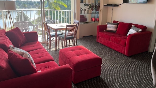 A living room with a red sofa, a red ottoman, a dining table with chairs, a TV mounted on the wall, and a balcony with a view of palm trees. - Hilo's Finest 2 bd Condo - Pool, A/C, Views! (Hilo)