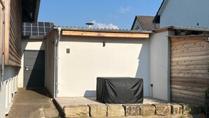 A covered outdoor seating area with a stone border, a solar panel on the roof, and a white building with a wooden fence. - Modern vacation apartment Knüllwald (Knüllwald)