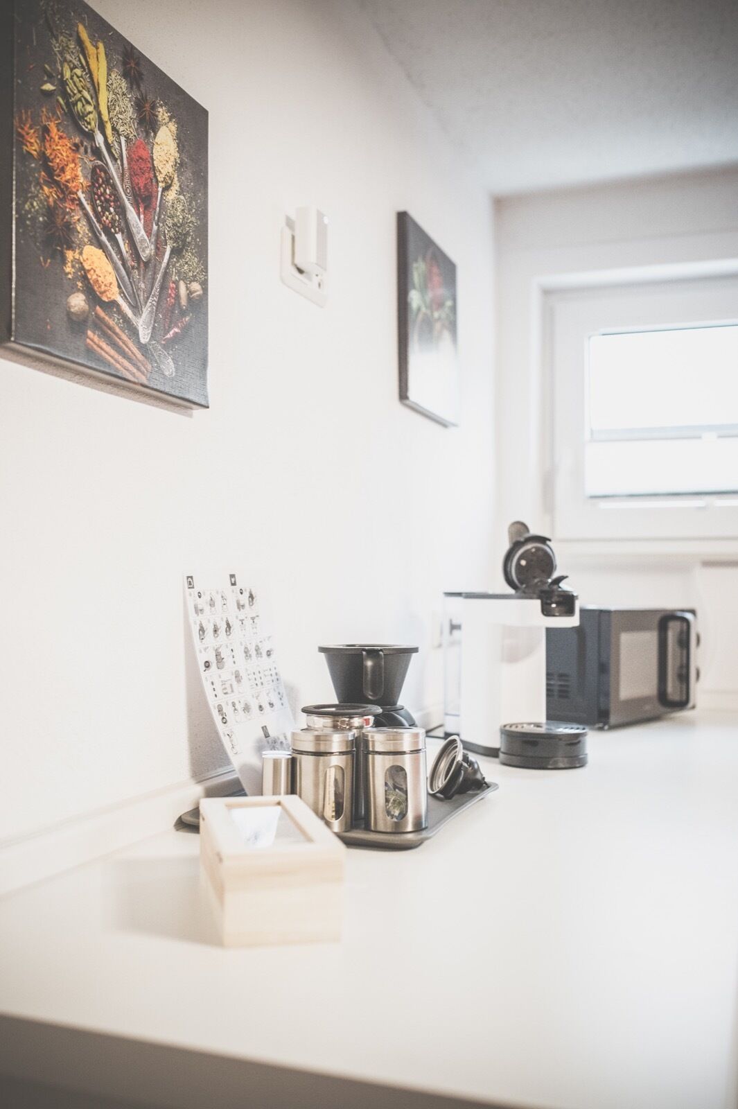 A kitchen counter with a coffee maker, a kettle, and a coffee grinder. There are spices and a recipe chart on the wall.