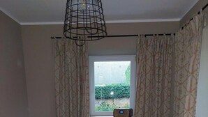 Una habitación acogedora con un sofá estampado de cuadros, una pequeña mesa de comedor con dos sillas blancas, una ventana con cortinas y un radio montado en la pared.