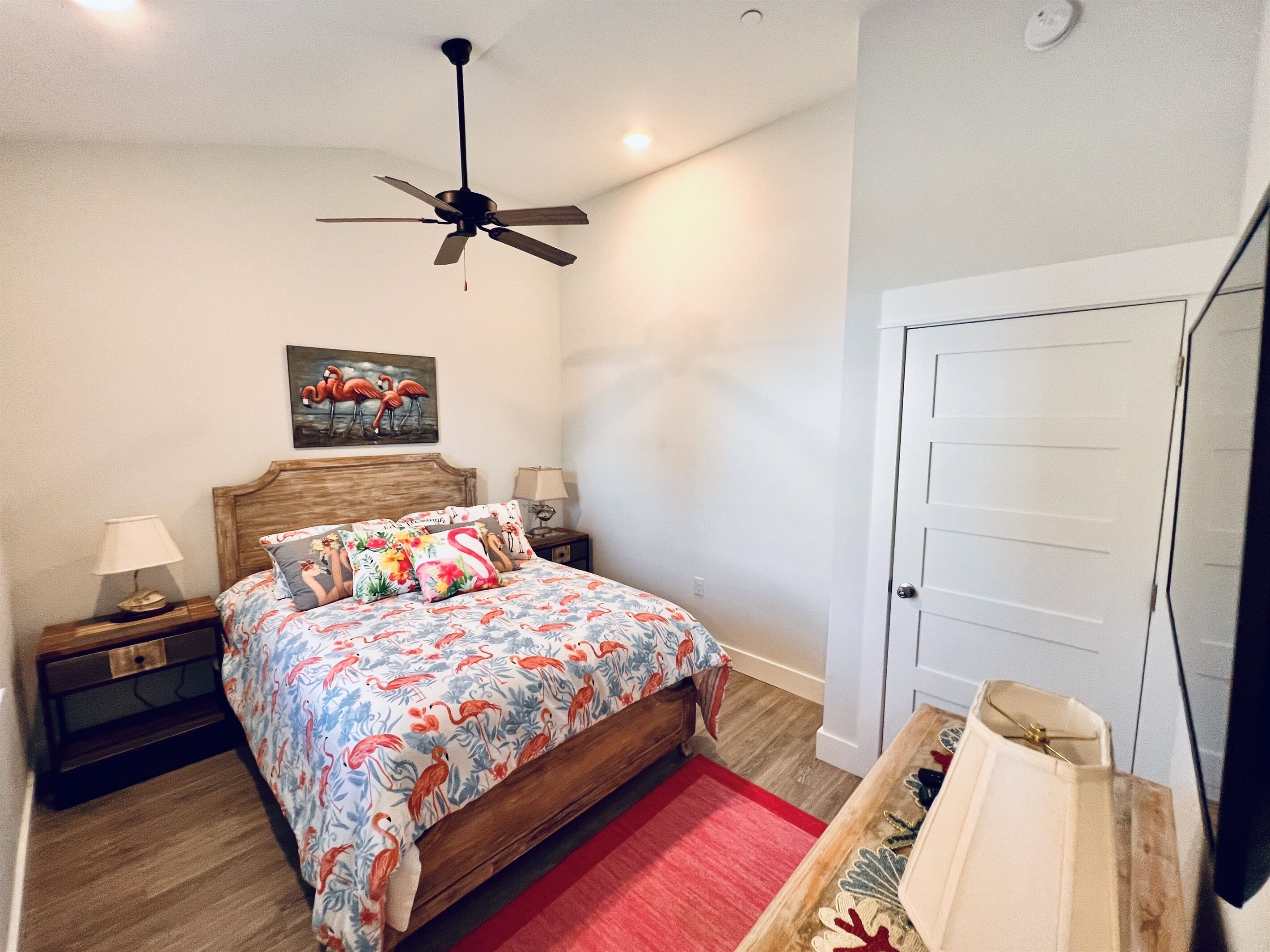 A bedroom with a wooden bed, a ceiling fan, a nightstand with a lamp, a red rug, and a door.