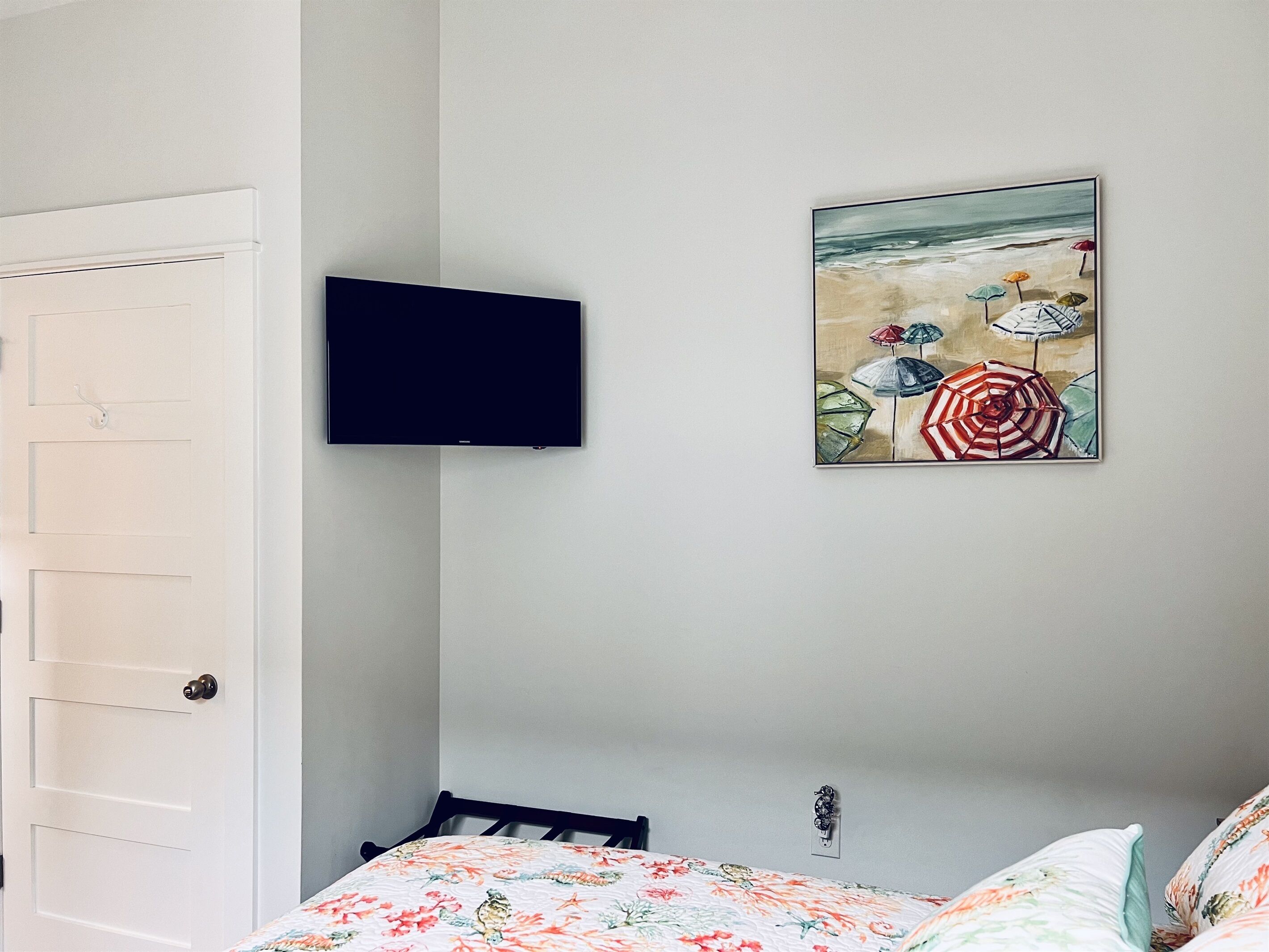 A bedroom with a bed, a television mounted on the wall, and a painting of beach umbrellas.